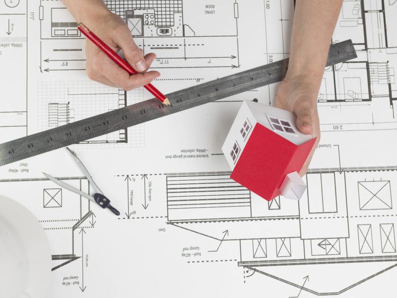 high-angle-view-hand-holding-small-house-model-pencil-blueprint-min